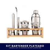 Set de coctelería plateado organizado en una base de madera. Incluye coctelera, medidor, cuchara mezcladora, macerador, colador y dosificadores sobre fondo blanco. En la parte inferior tiene un banner azul con el texto "Kit Bartender Plateado Llévalo con tu botella de Ron preferida".
