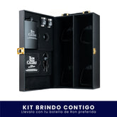 Elegante estuche negro abierto de Ron Viejo de Caldas. En el lado izquierdo incluye una licorera de bolsillo negra, dos vasos metálicos tipo shot, un embudo y accesorios con el logo de la marca. El lado derecho tiene compartimentos vacíos para botellas, todo sobre fondo blanco. En la parte inferior tiene un banner azul con el texto "Kit Brindo Contigo Llévalo con tu botella de Ron preferida"