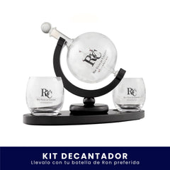 Set de bar que incluye un decantador de vidrio esférico montado sobre un soporte giratorio y dos vasos de cristal, todos con el logo de Ron Viejo de Caldas grabado, sobre fondo blanco. En la parte inferior tiene un banner azul con el texto "Kit Decantador Llévalo con tu botella de Ron preferida".