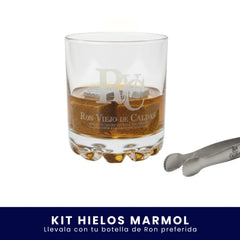 Vaso de cristal grabado con el logo de Ron Viejo de Caldas, servido con licor y cubos enfriadores de mármol en su interior, junto a unas pinzas metálicas sobre fondo blanco. En la parte inferior tiene un banner azul con el texto "Kit Hielos Mármol Llévala con tu botella de Ron preferida".